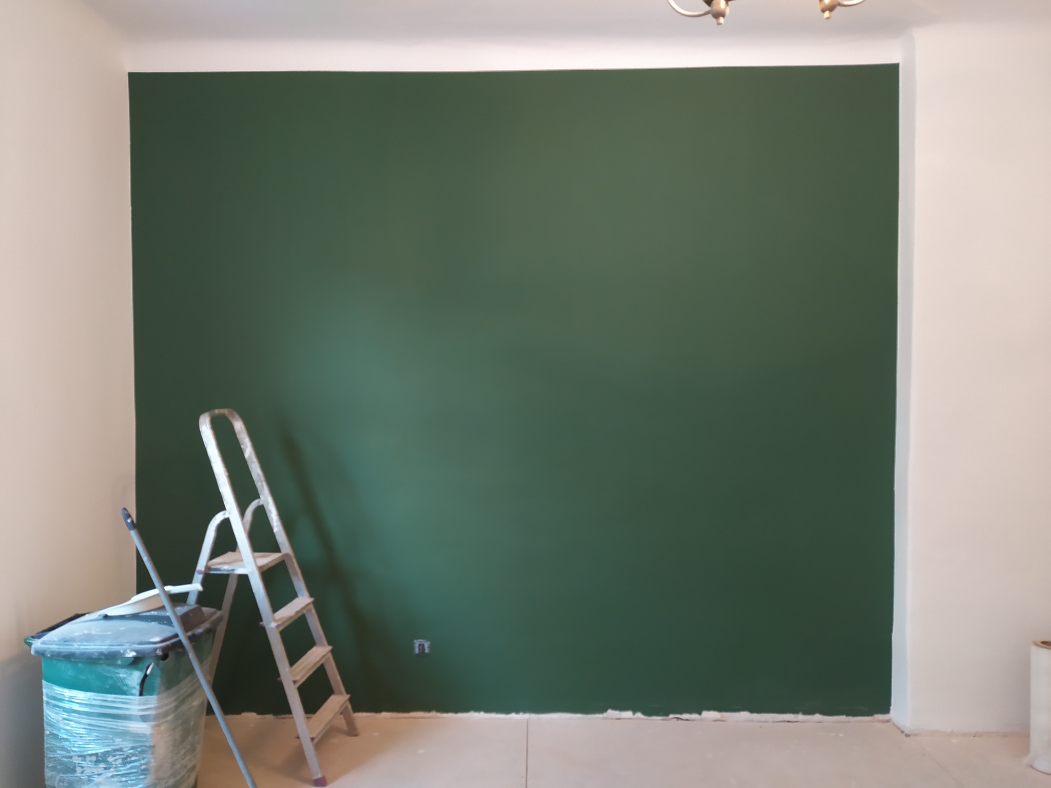 Świeżo pomalowana na ciemnozielono ściana w pokoju, z aluminiową drabiną i wiadrem z farbą obok, widoczny fragment gniazdka elektrycznego na ścianie.