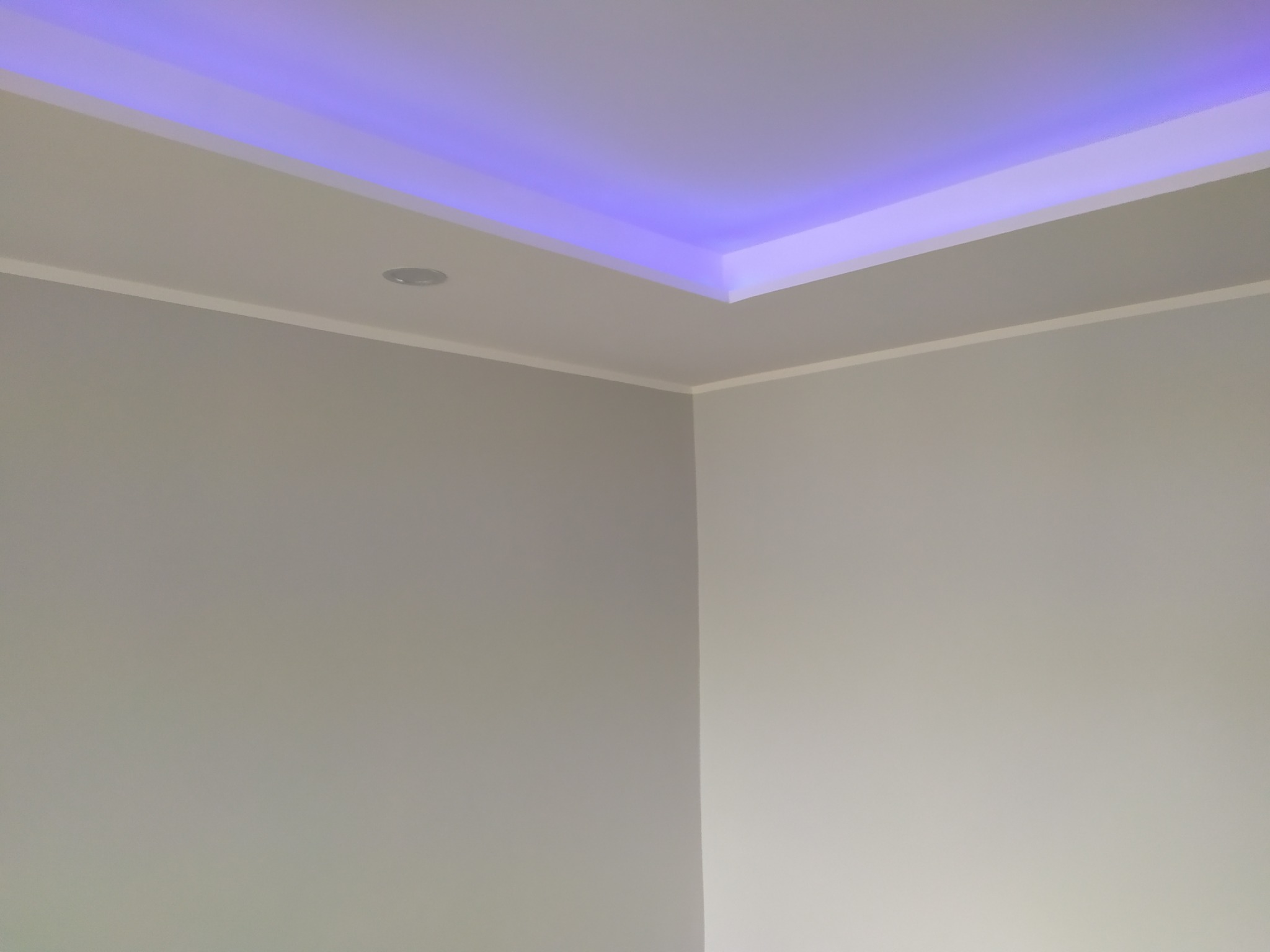 Wykończony róg pokoju z pomalowanymi ścianami w odcieniach szarości i białym sufitem z podwieszanym oświetleniem LED o barwie fioletowej.