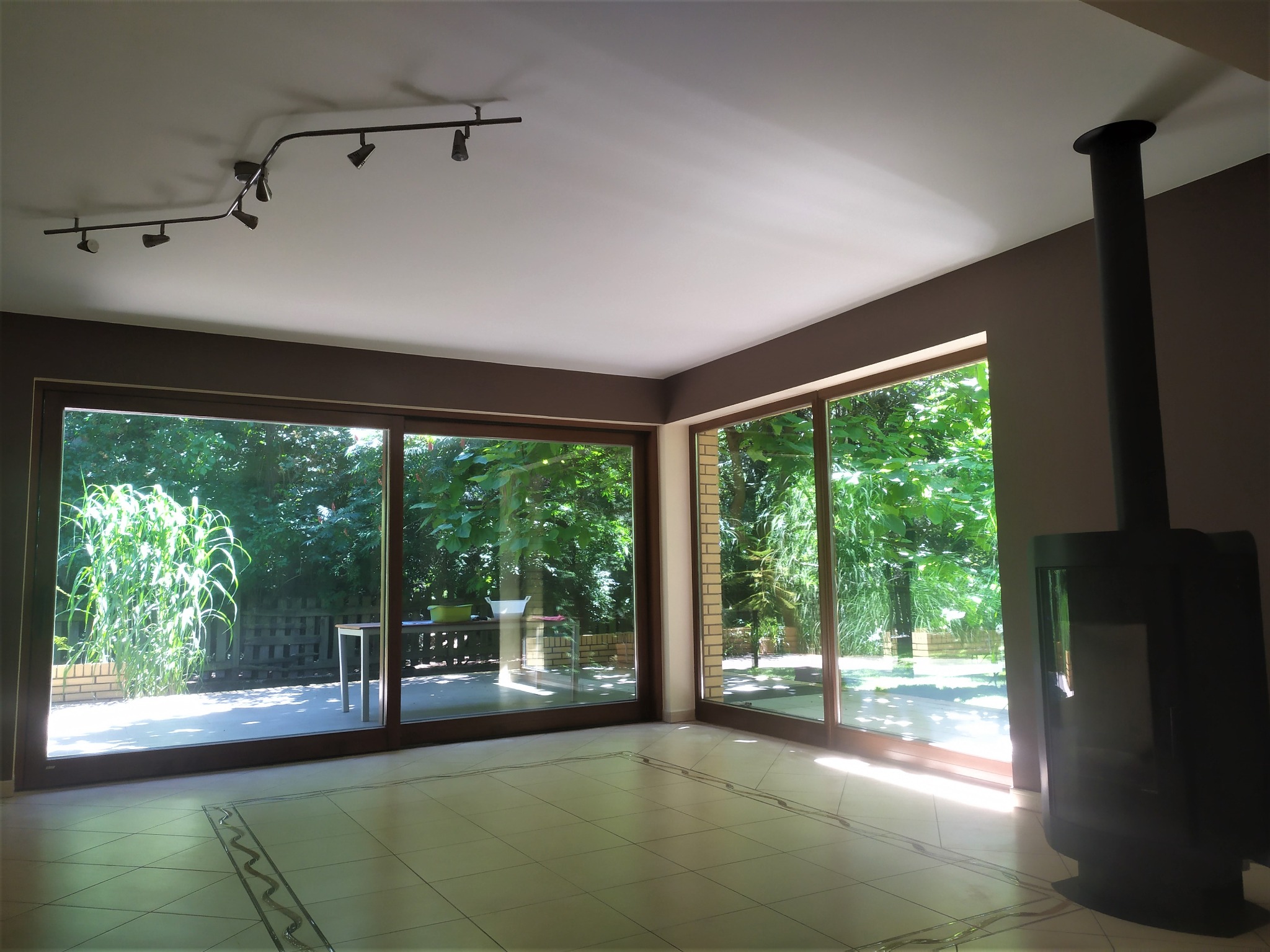 Jasne, przestronne wnętrze z dużymi przesuwnymi oknami wychodzącymi na ogród, ciemnobrązowe ramy okienne i listwy przypodłogowe, kremowe płytki podłogowe z dekoracyjnym wzorem, nowoczesny czarny...