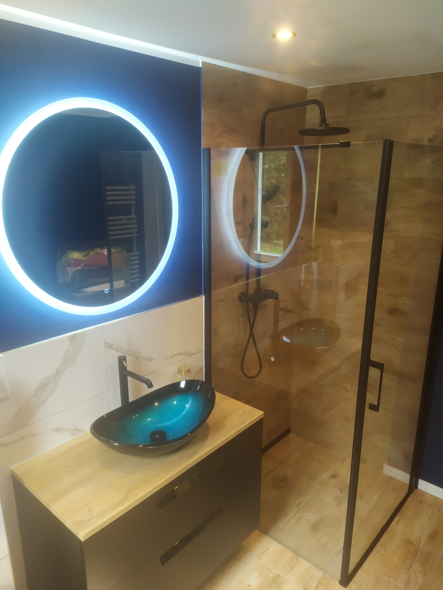 Nowoczesna łazienka z kabiną prysznicową typu walk-in, umywalką nablatową w odcieniach błękitu i czarną armaturą, oświetlona okrągłym lustrem LED.