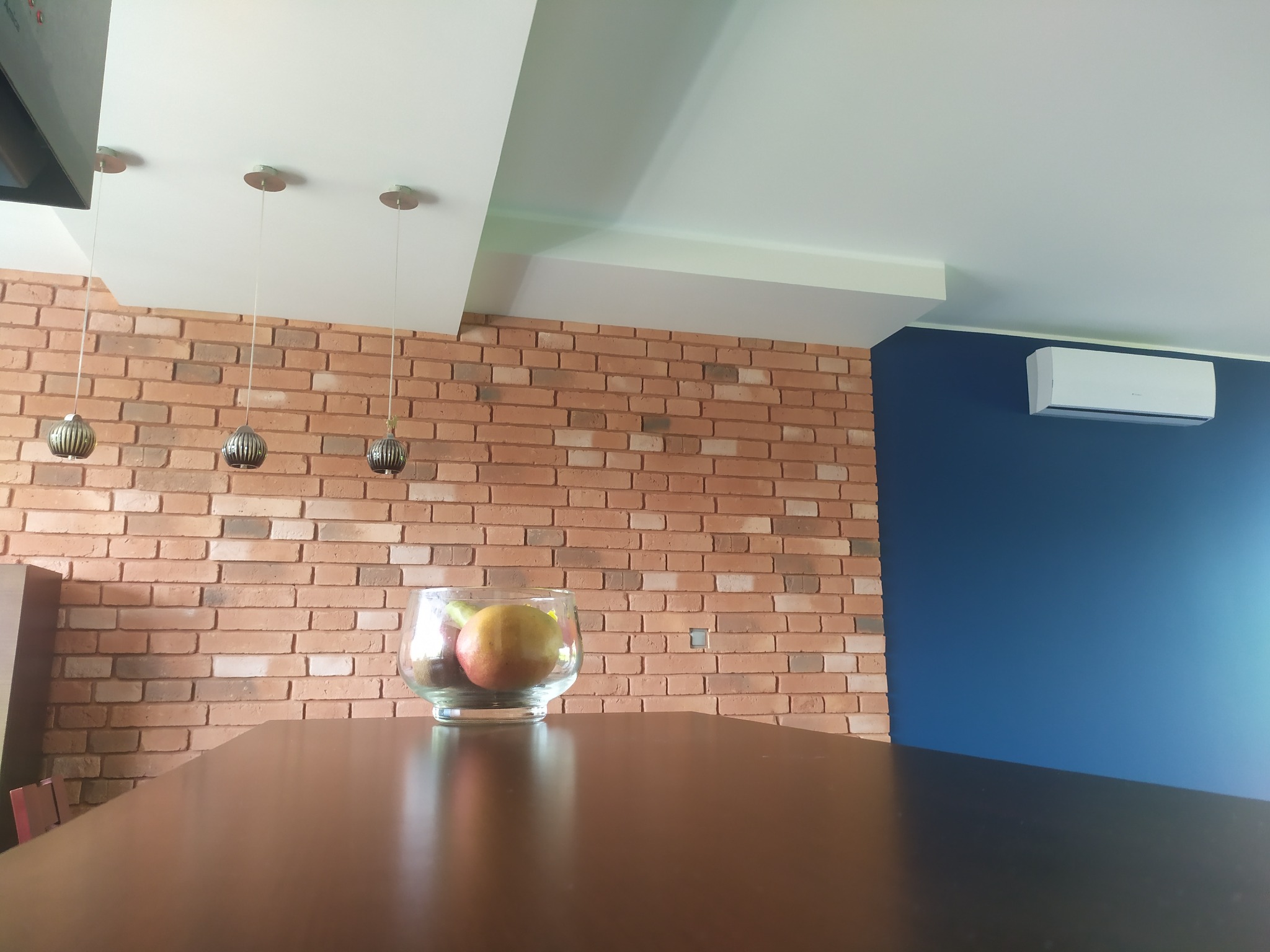 Wnętrze z ceglaną ścianą, trzema wiszącymi lampami, owocami w szklanym wazonie na blacie i niebieską ścianą z klimatyzatorem.
