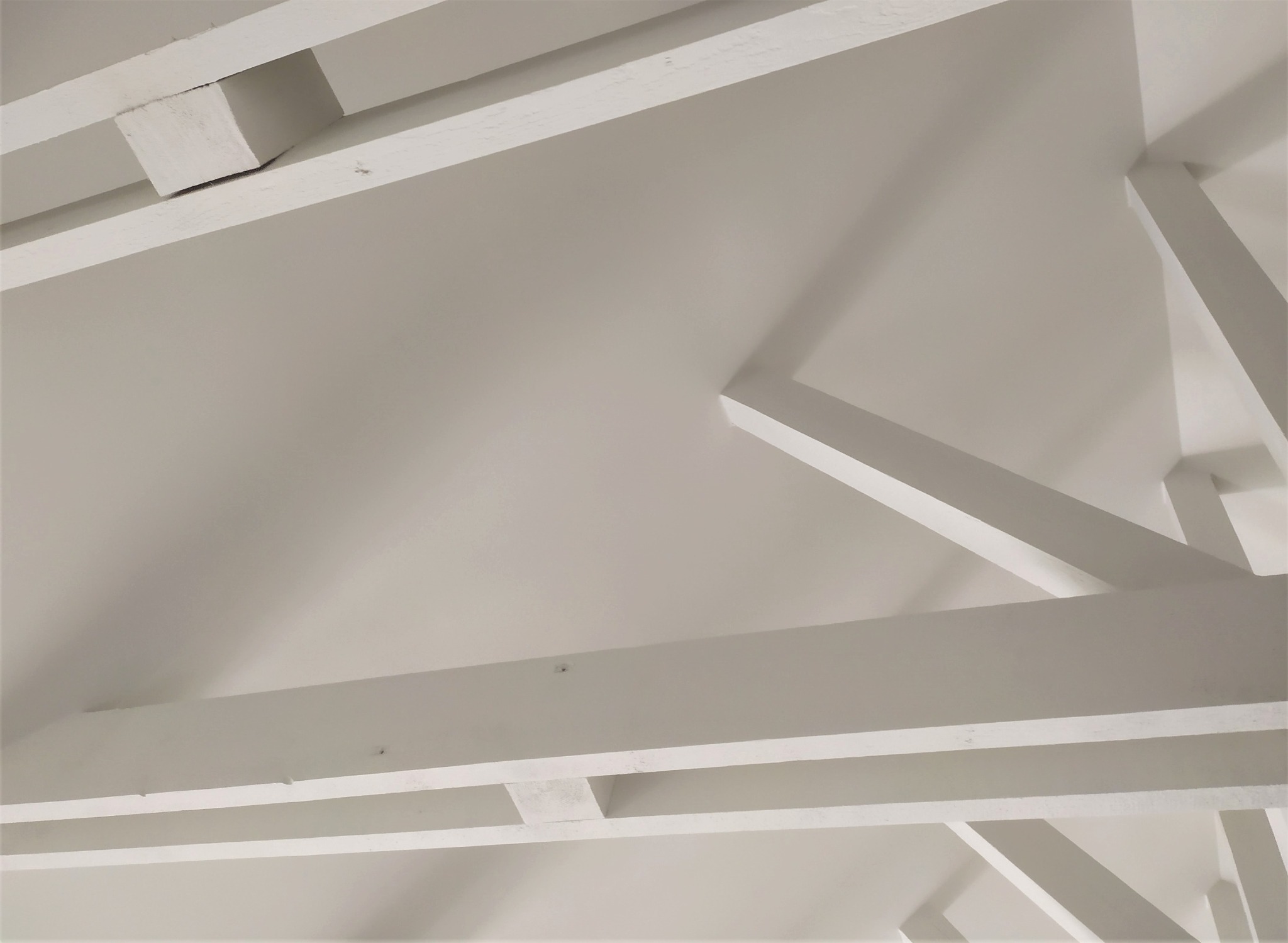 Biały sufit z drewnianymi belkami konstrukcyjnymi, widoczne ślady po szpachlowaniu i malowaniu, kąt patrzenia od dołu.