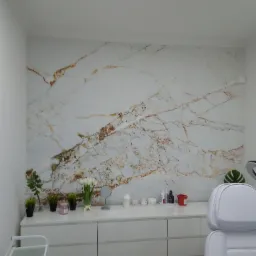 Ściana wykończona płytą imitującą marmur z brązowymi żyłami w gabinecie kosmetycznym, biała komoda z dekoracjami i fotel kosmetyczny w tle.