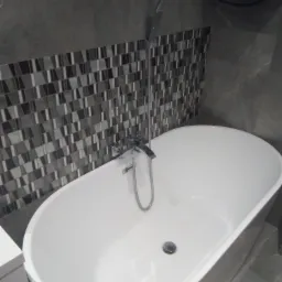 Nowoczesna, wolnostojąca wanna owalna z chromowaną armaturą na tle ściany wyłożonej mozaiką w odcieniach szarości i czerni.