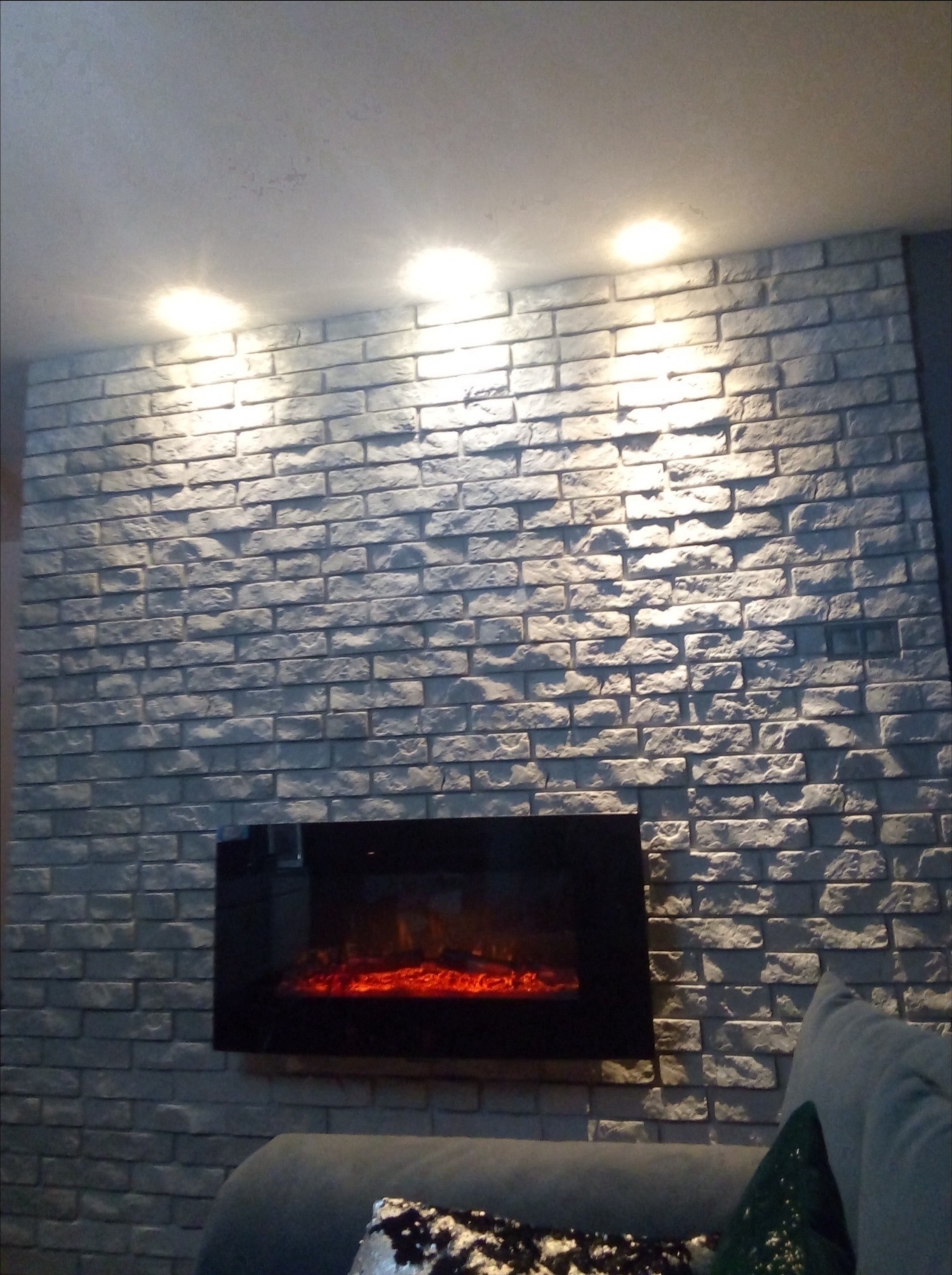 Ściana wykończona dekoracyjną szarą cegłą z zamontowanym elektrycznym kominkiem z efektem płomieni, oświetlona punktowymi lampami sufitowymi.