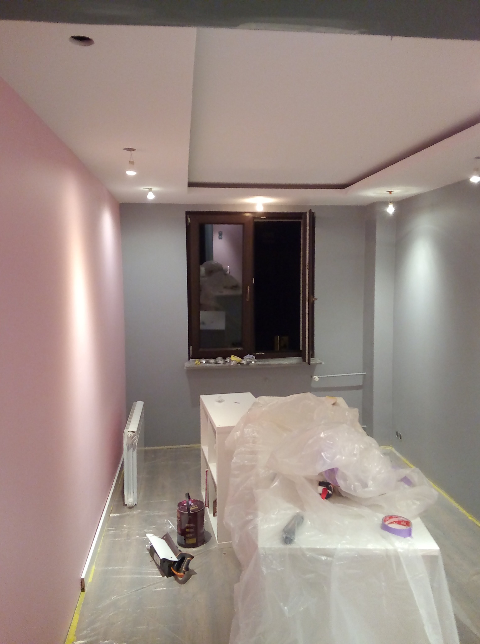 Pokój w trakcie odświeżania: jedna ściana pomalowana na różowo, druga na szaro, okno z drewnianą ramą, podłoga zabezpieczona folią, widoczne puszki z farbą i narzędzia malarskie.