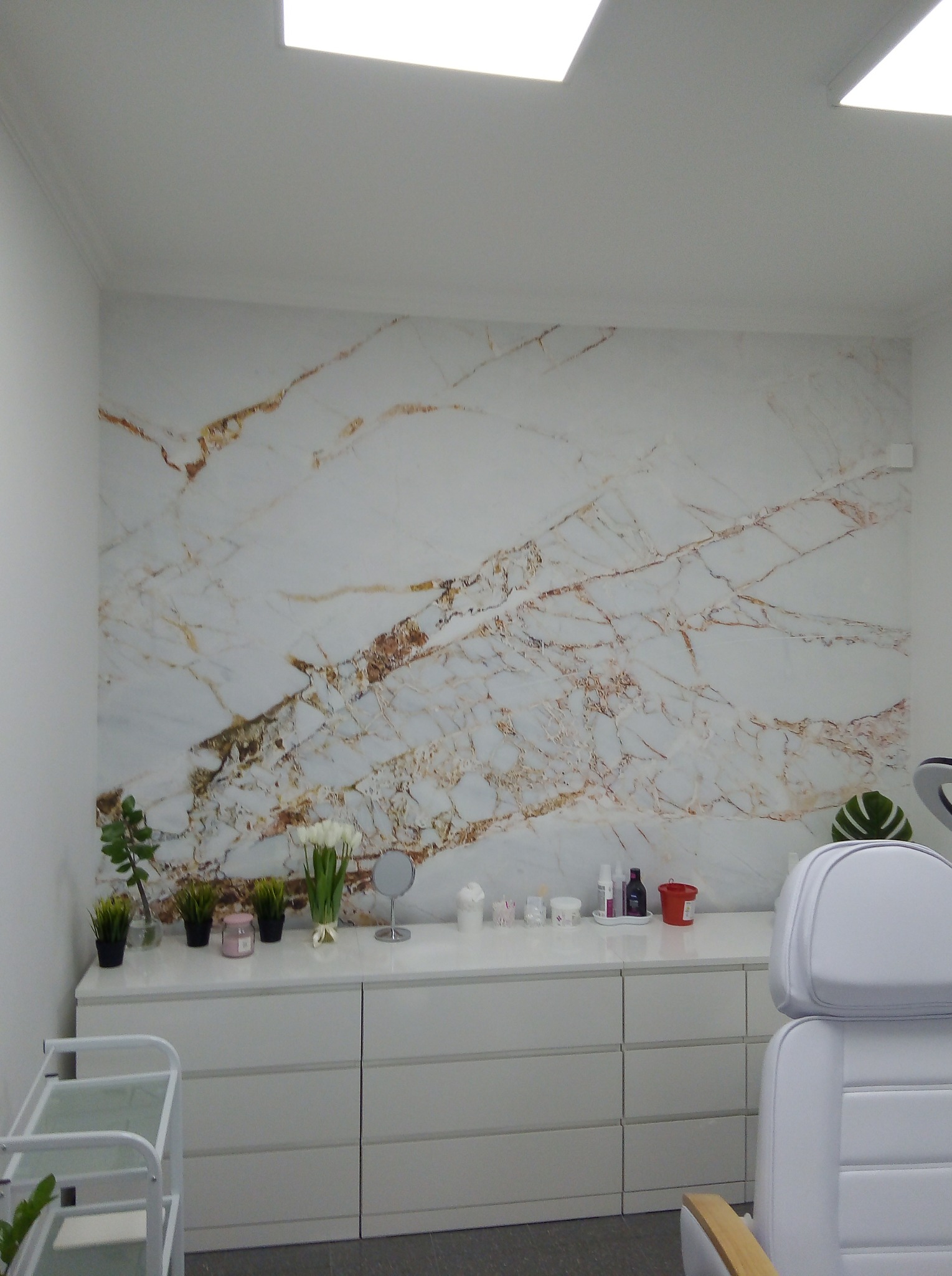 Ściana wykończona płytą imitującą marmur z brązowymi żyłami w gabinecie kosmetycznym, biała komoda z dekoracjami i fotel kosmetyczny w tle.