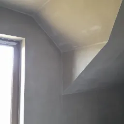 Świeżo nałożony tynk maszynowy na skośnej ścianie poddasza, widoczne okno z naklejkami producenta DAKO oraz sufit z lekkimi niedoskonałościami wykończenia.