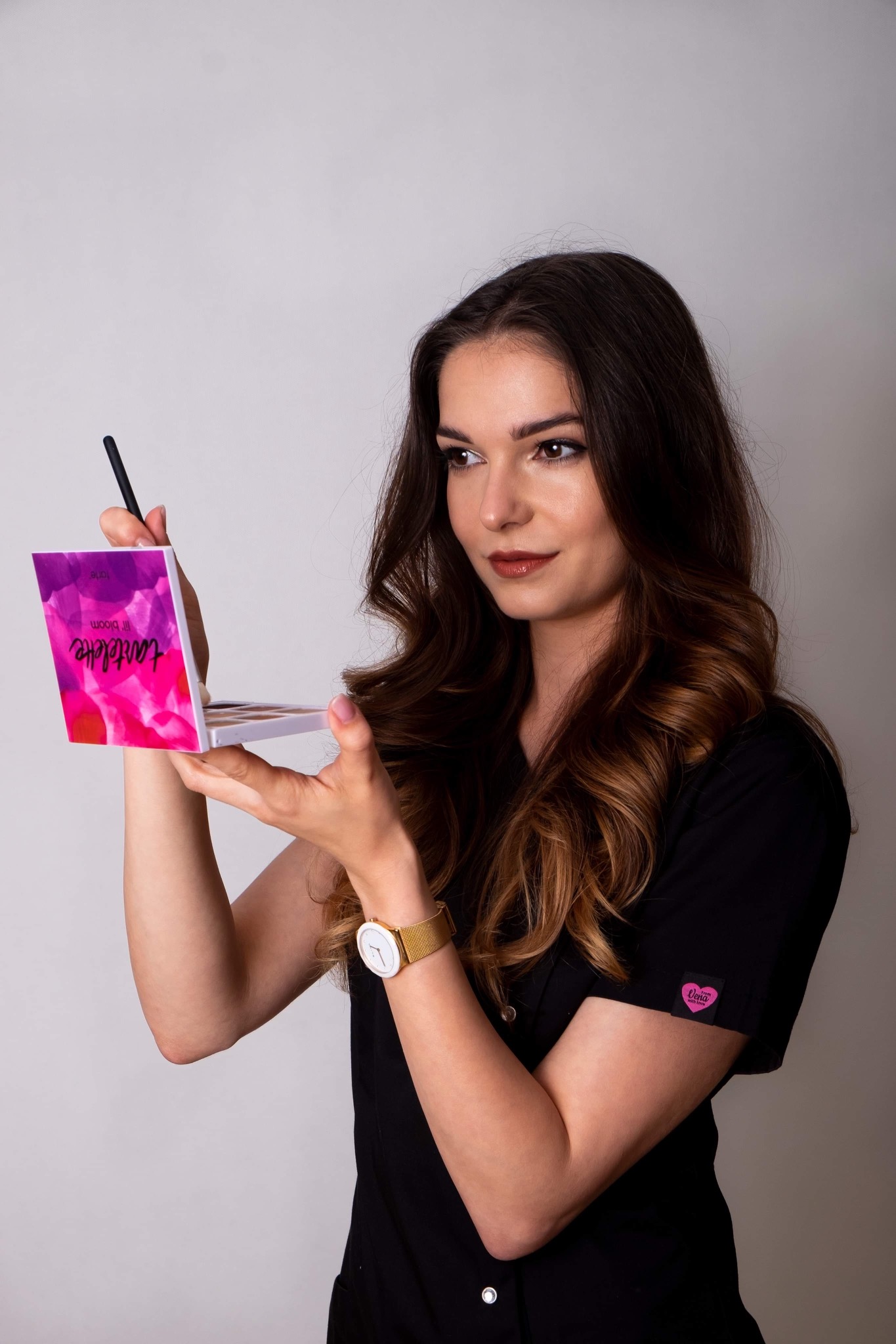 Kobieta w czarnym uniformie trzyma paletę cieni Tartelette Lil Bloom i pędzel do makijażu, spoglądając w bok.