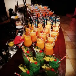 Bar z rzędami kolorowych drinków: niebieskie, pomarańczowe i zielone, każdy ozdobiony owocami i słomkami, przygotowane na przyjęcie.