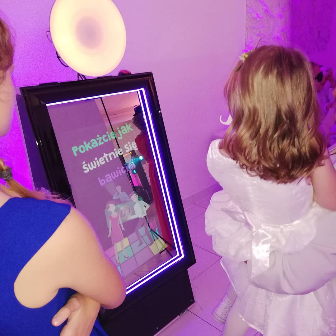 Dziewczynka w białej sukience stoi przed interaktywnym lustrem z napisem 'Pokażcie jak świetnie się bawicie' i animacjami postaci na parkiecie, oświetlonym fioletowym światłem.