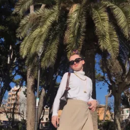 Kobieta w okularach przeciwsłonecznych, białym golfie i beżowej spódnicy na tle palmy i miejskiego krajobrazu.