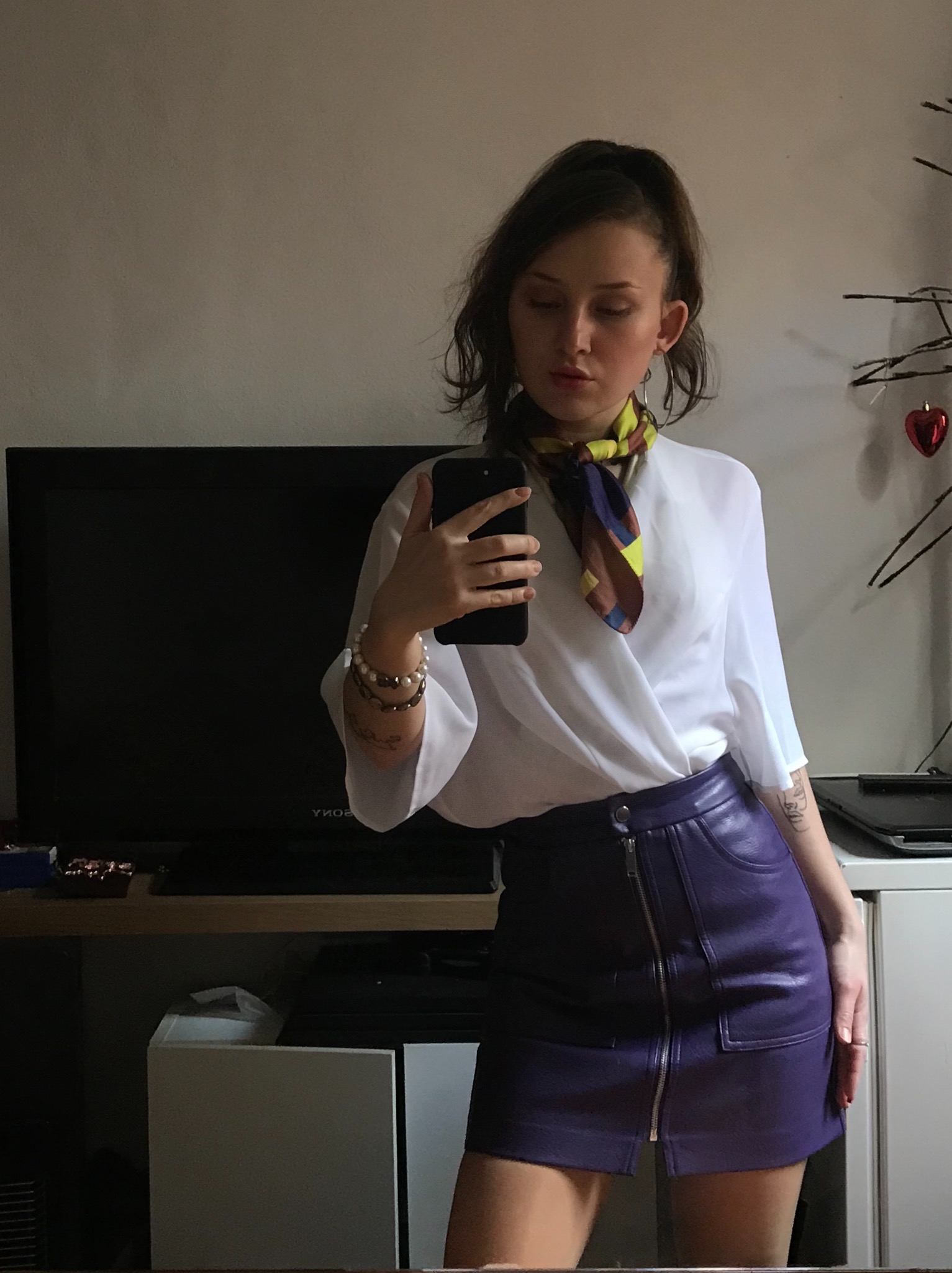 Kobieta robi selfie w lustrze, ubrana w białą bluzkę, fioletową skórzaną spódnicę i wzorzystą apaszkę.