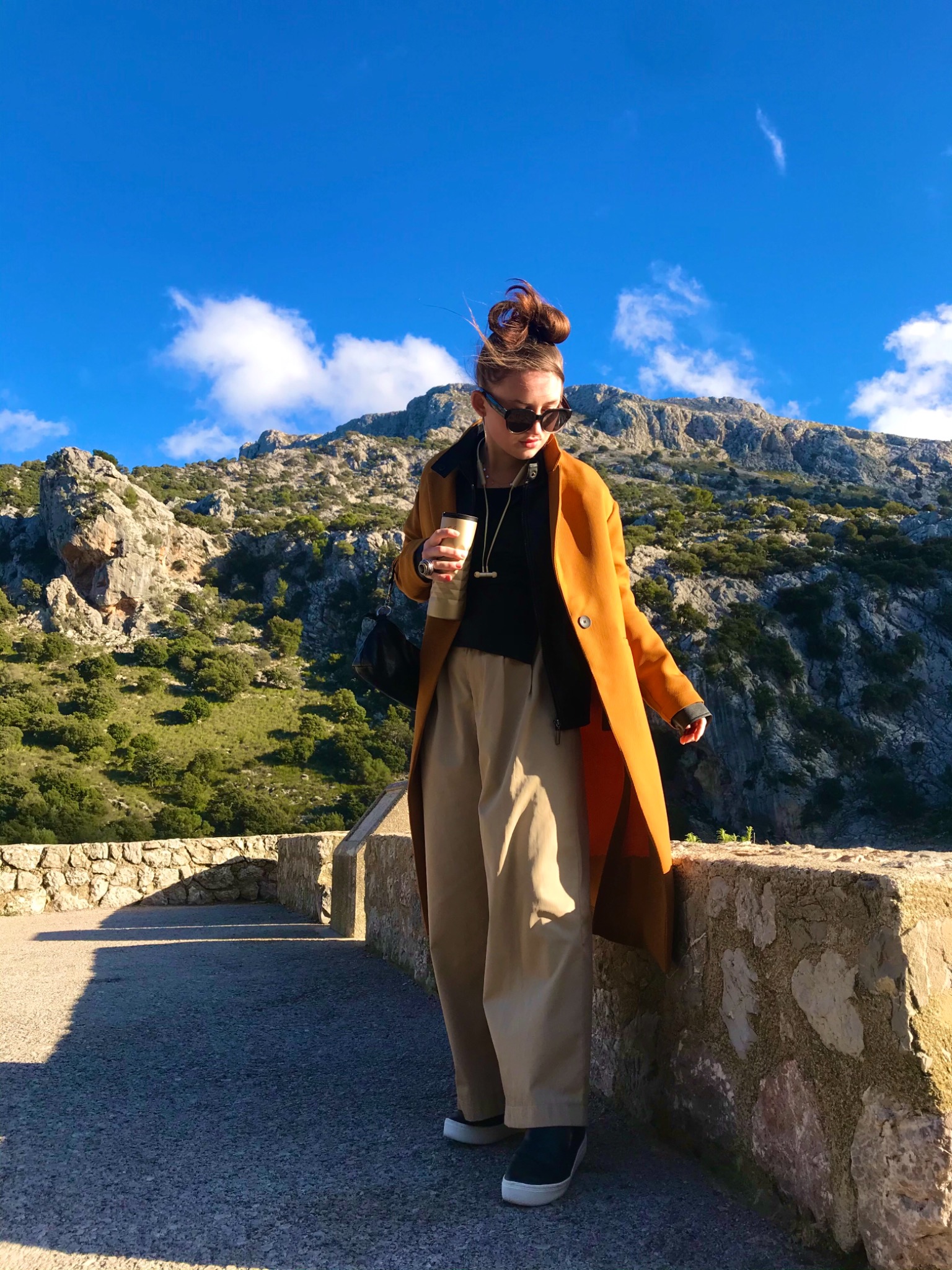 Kobieta w pomarańczowym płaszczu, szerokich spodniach i okularach przeciwsłonecznych, stojąca na kamiennym murku z kubkiem termicznym w ręku, w tle góry i błękitne niebo.