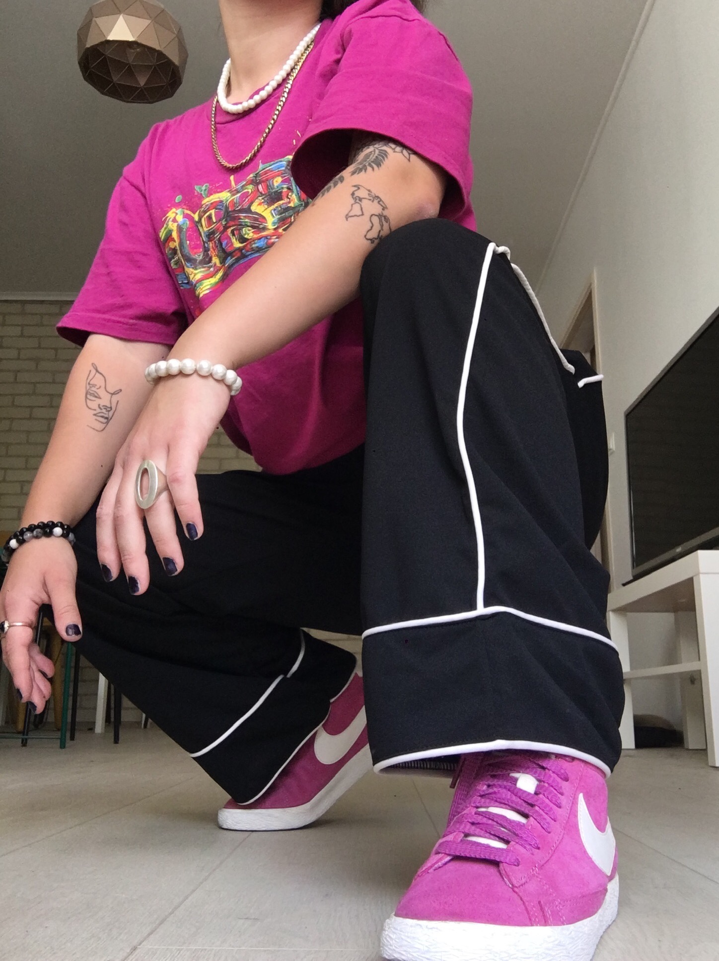 Osoba w kucki prezentuje stylizację: różowa koszulka z nadrukiem, czarne spodnie z białym lampasem, różowe sneakersy Nike, liczne tatuaże na ramionach, perłowe i kamienne bransoletki oraz ciemny...