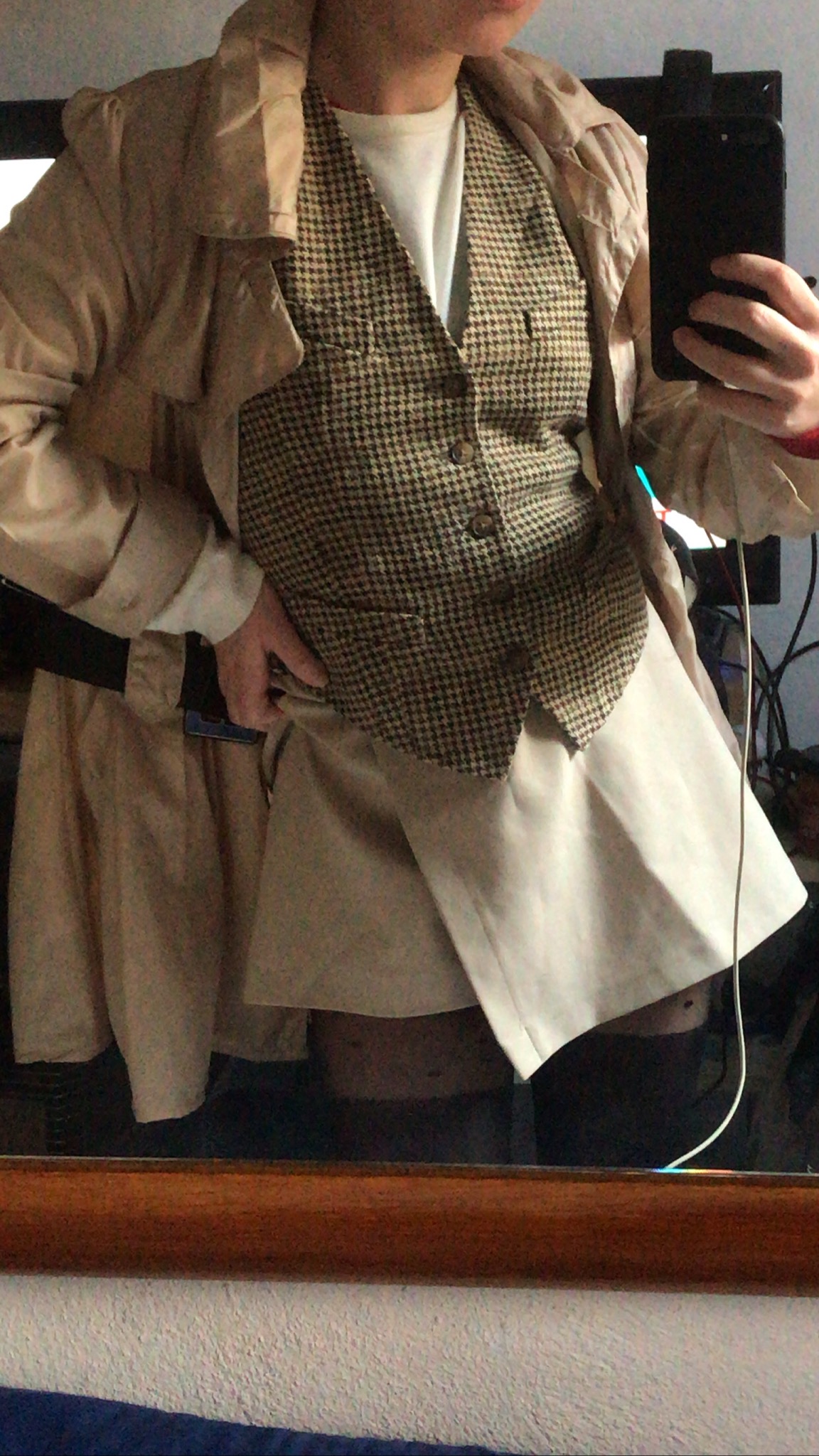 Osoba w beżowym płaszczu, kraciastej kamizelce i białej spódnicy pozuje do zdjęcia w lustrze.