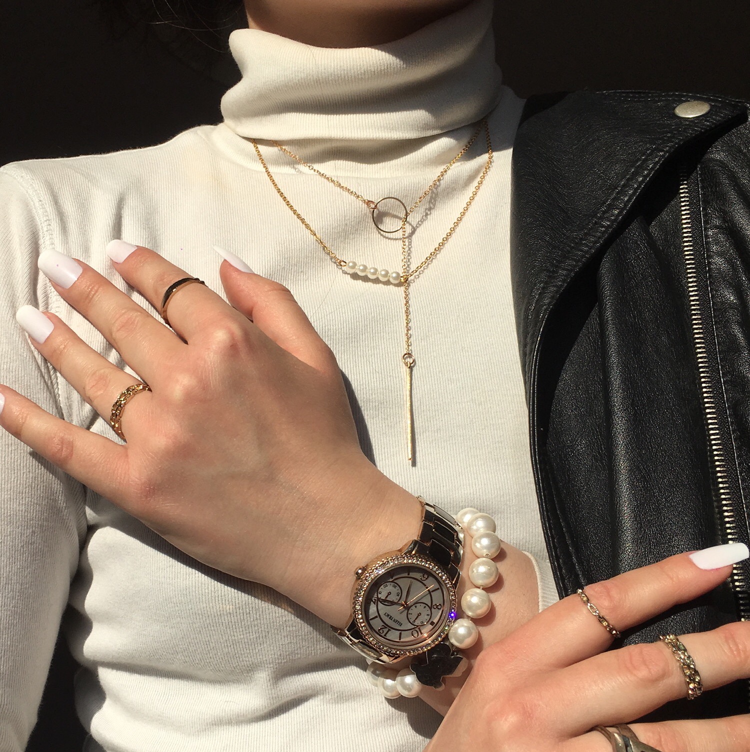 Kobieta w białym golfie prezentuje białe, kwadratowe paznokcie, złotą biżuterię: naszyjniki, pierścionki, zegarek i bransoletkę z pereł oraz czarną skórzaną kurtkę.