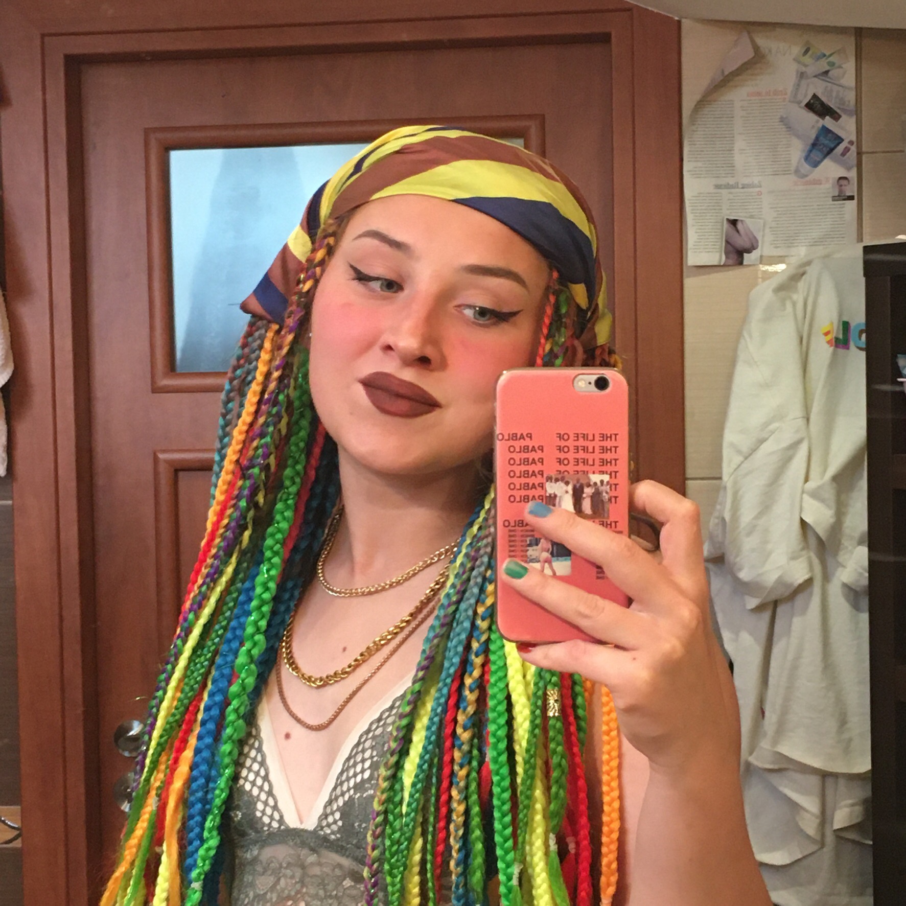 Selfie kobiety z kolorowymi warkoczykami, ciemną szminką i wyrazistym makijażem oczu, w chustce na głowie, pozującej w łazience.