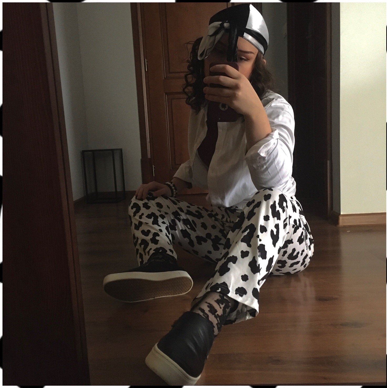Osoba siedząca na podłodze w spodniach w cętki, białej koszuli i czarno-białej czapce, robiąca selfie w lustrze.