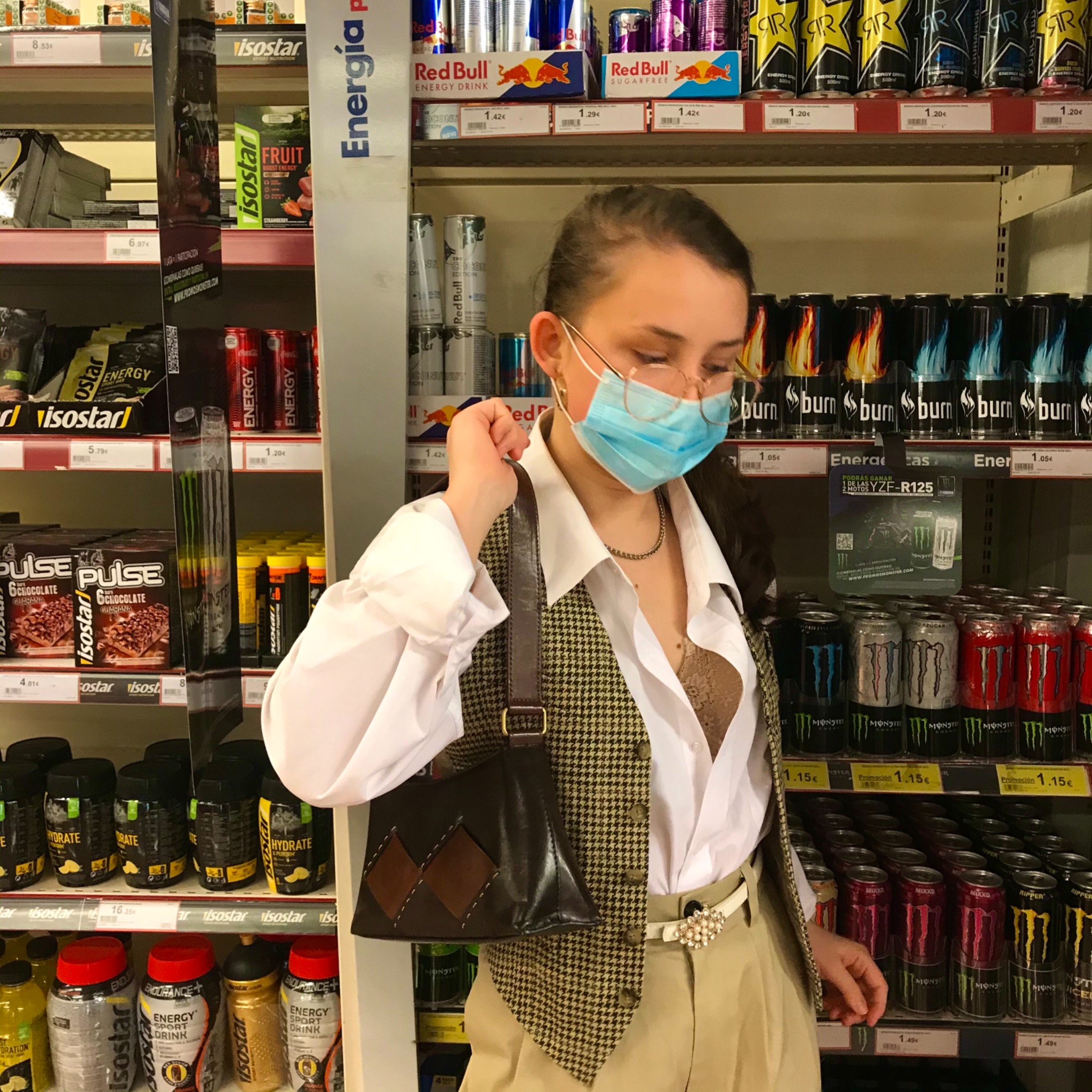 Osoba w masce, kamizelce w pepitkę i z torebką na ramieniu, robiąca zakupy w sklepie z napojami energetycznymi.