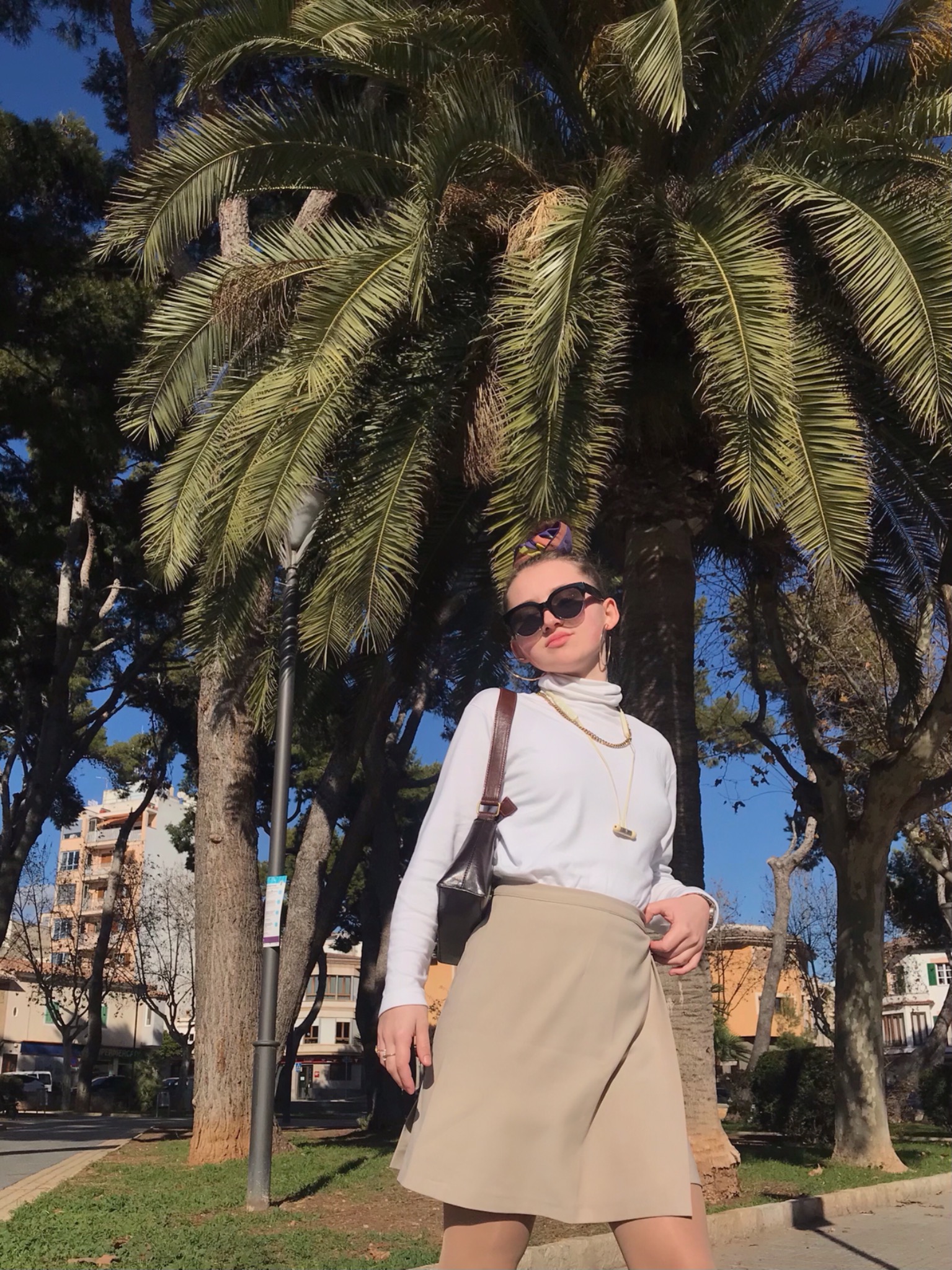 Kobieta w okularach przeciwsłonecznych, białym golfie i beżowej spódnicy na tle palmy i miejskiego krajobrazu.