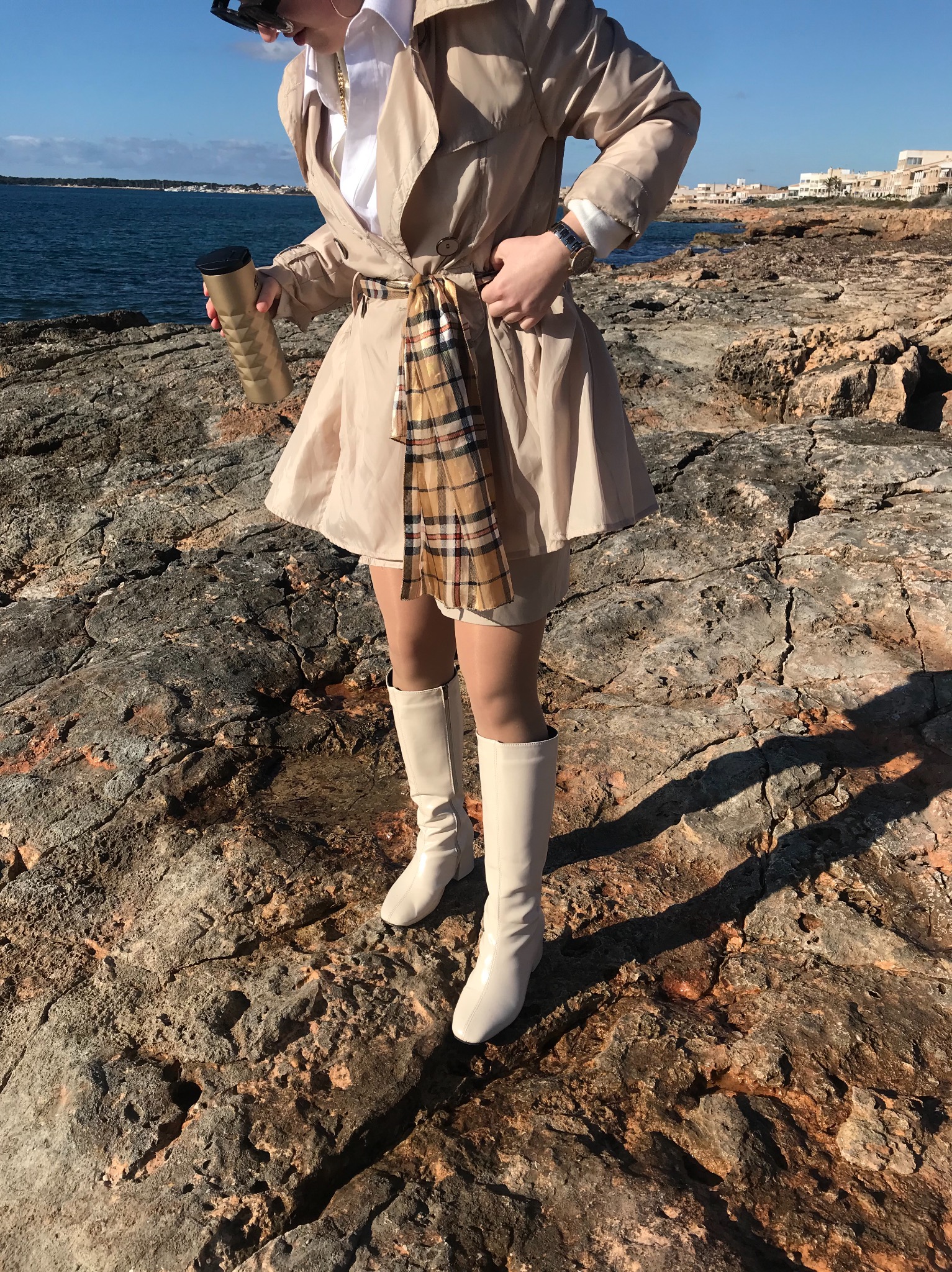 Kobieta w beżowym płaszczu, spódnicy i białych kozakach pozuje na skalistym wybrzeżu z widokiem na morze i budynki w oddali, trzymając złotą butelkę termiczną.