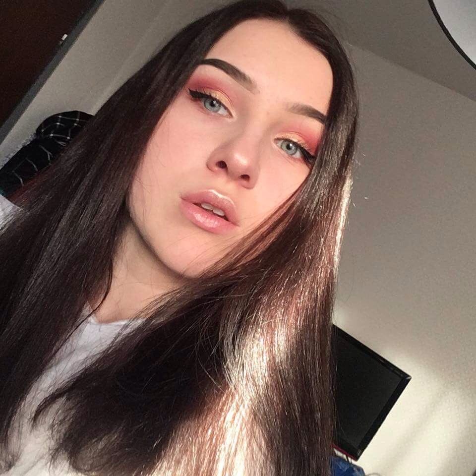 Portret młodej kobiety z intensywnym makijażem oczu w odcieniach różu i brzoskwini z wyraźną kreską oraz błyszczącymi ustami, uchwycony w naturalnym świetle.