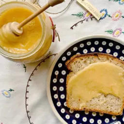 Słoik z jasnym, kremowym produktem z zanurzoną drewnianą nabieraczką, obok kromka chleba posmarowana tym samym produktem na talerzu w stylu Bolesławiec, całość na haftowanym obrusie z etykietą...