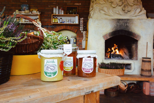 Trzy słoiki z różnymi rodzajami miodu na drewnianym stole, w tle kominek z palącym się ogniem oraz kosze z roślinami i ziołami.