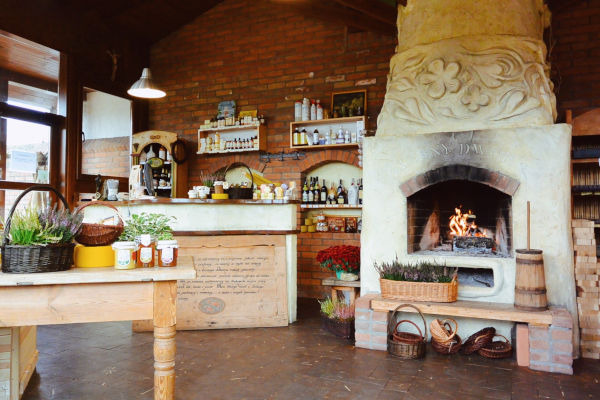 Rustykalne wnętrze sklepu w Szczytnie z ceglaną ścianą, półkami z produktami, kominkiem i stołem z koszykami i słoikami z produktami.
