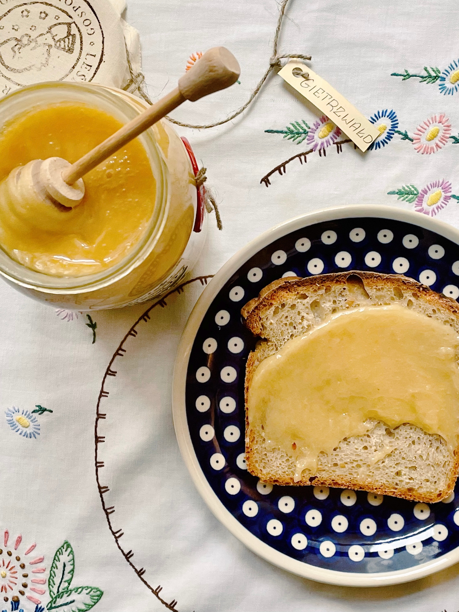 Słoik z jasnym, kremowym produktem z zanurzoną drewnianą nabieraczką, obok kromka chleba posmarowana tym samym produktem na talerzu w stylu Bolesławiec, całość na haftowanym obrusie z etykietą...