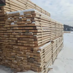 Stos tarcicy budowlanej pokryty świeżym śniegiem na placu składowym w zimowy dzień.