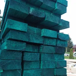 Stos impregnowanych na zielono kantówek drewnianych ułożonych równo, widoczne słoje drewna i struktura impregnowanej powierzchni, w tle zieleń i budynki.