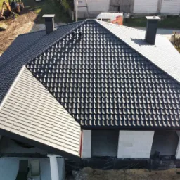 Widok z góry na dom w stanie surowym zamkniętym z nowym, ciemnoszarym pokryciem dachowym z dachówki modułowej i dwoma kominami. W tle widoczne inne budynki i plac budowy.
