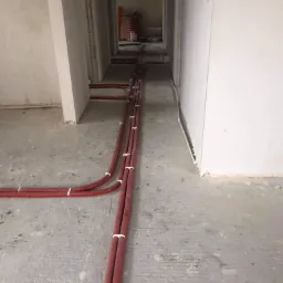 Czerwone rury ogrzewania podłogowego, przymocowane białymi opaskami, ułożone na surowym betonowym podłożu w trakcie instalacji w nowym budynku.