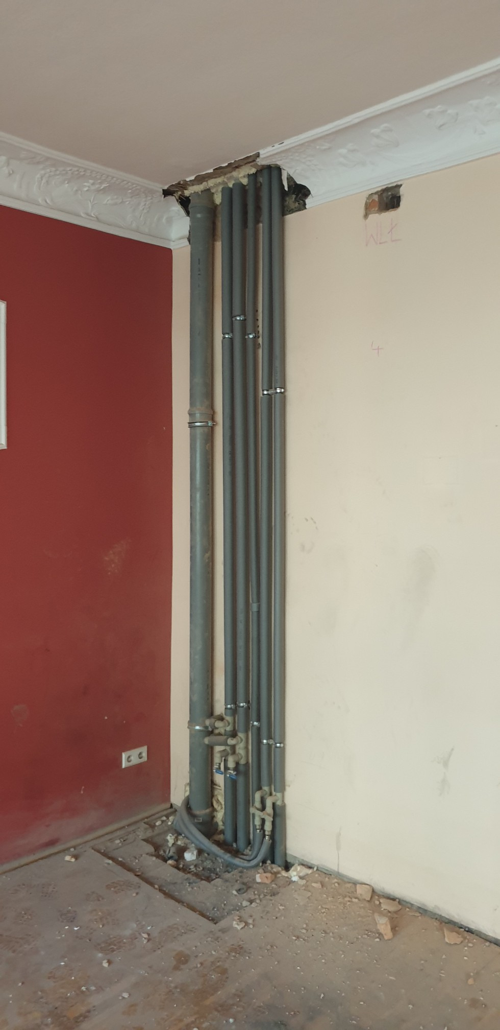 Szare rury grzewcze, prowadzone wzdłuż ściany, z widocznymi zaworami i mocowaniami, wychodzące z dziury w suficie, otoczone pianką montażową, w pomieszczeniu w trakcie remontu.