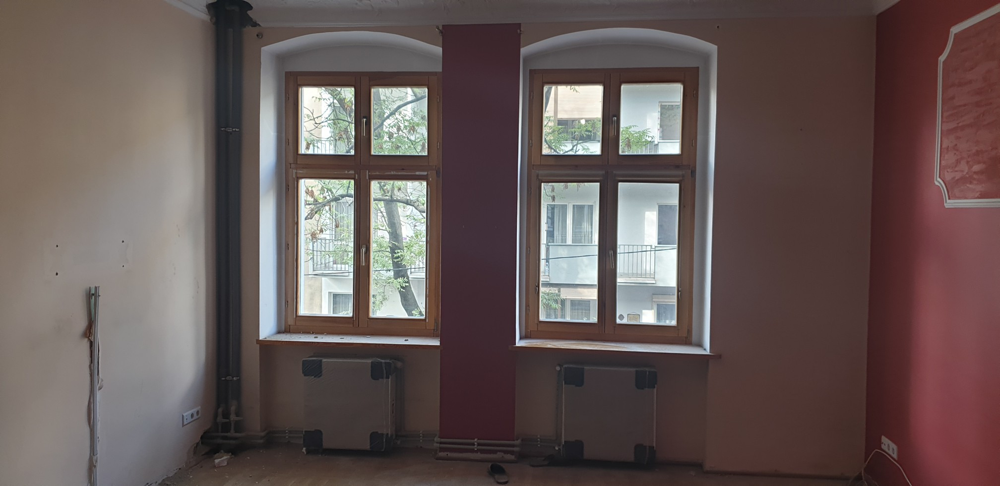 Pokój przed remontem z dwoma oknami w drewnianych ramach, pod którymi znajdują się grzejniki zabezpieczone folią. Widoczne pionowe rury grzewcze przy ścianie.