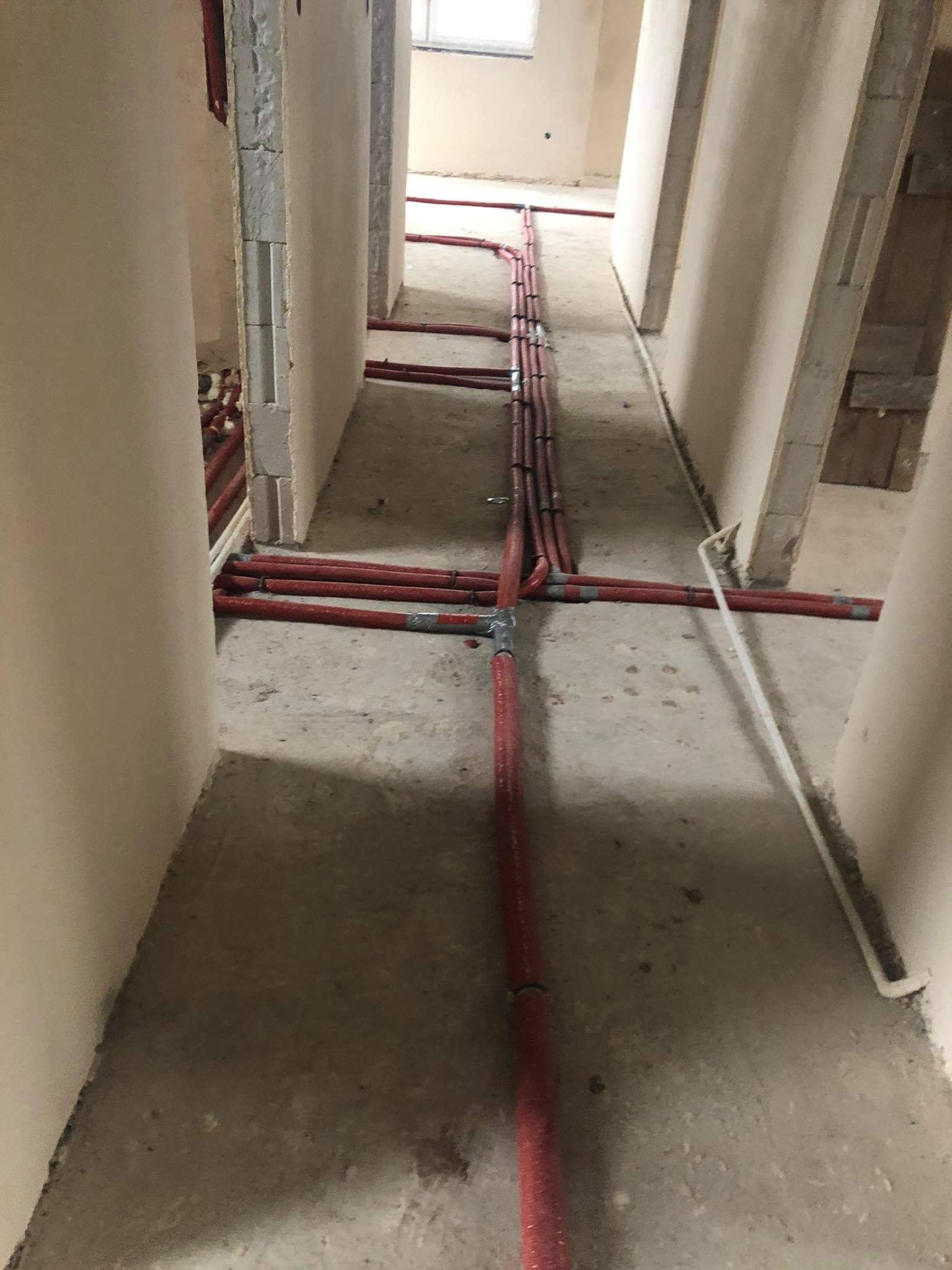 Czerwone rury rozprowadzone po podłodze w surowym stanie, widok wzdłuż korytarza z niedokończonymi ścianami.