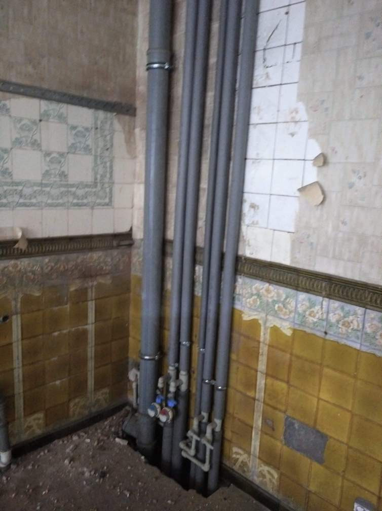 Szare rury pionowe z zaworami na tle zniszczonych, żółtych i białych płytek w starym budynku.