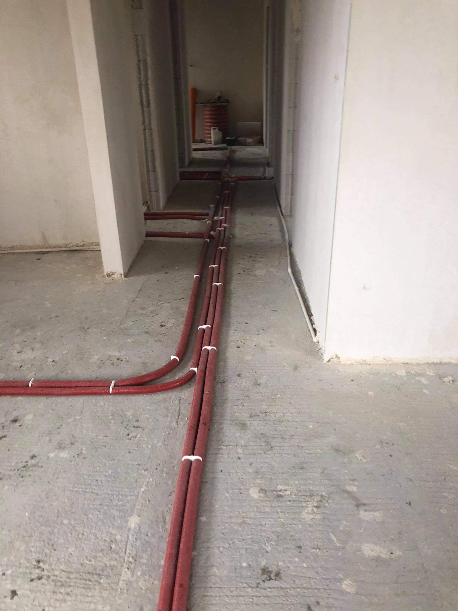 Czerwone rury ogrzewania podłogowego, przymocowane białymi opaskami, ułożone na surowym betonowym podłożu w trakcie instalacji w nowym budynku.