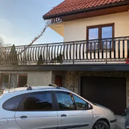 Czarna metalowa balustrada balkonowa z motywem greckim na tle beżowego domu z czerwoną dachówką, widok z poziomu zaparkowanego srebrnego samochodu.