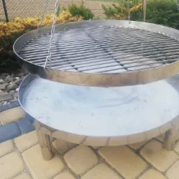 Okrągły, dwupoziomowy grill ze stali nierdzewnej z rusztem wiszącym na łańcuchach, ustawiony na kostce brukowej w ogrodzie z krzewami w tle.