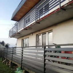 Widok z dołu na dwukondygnacyjny dom z balkonami zabezpieczonymi poziomymi, metalowymi barierkami. Na parterze widoczne ogrodzenie z poziomych paneli, częściowo zasłaniające rowery.