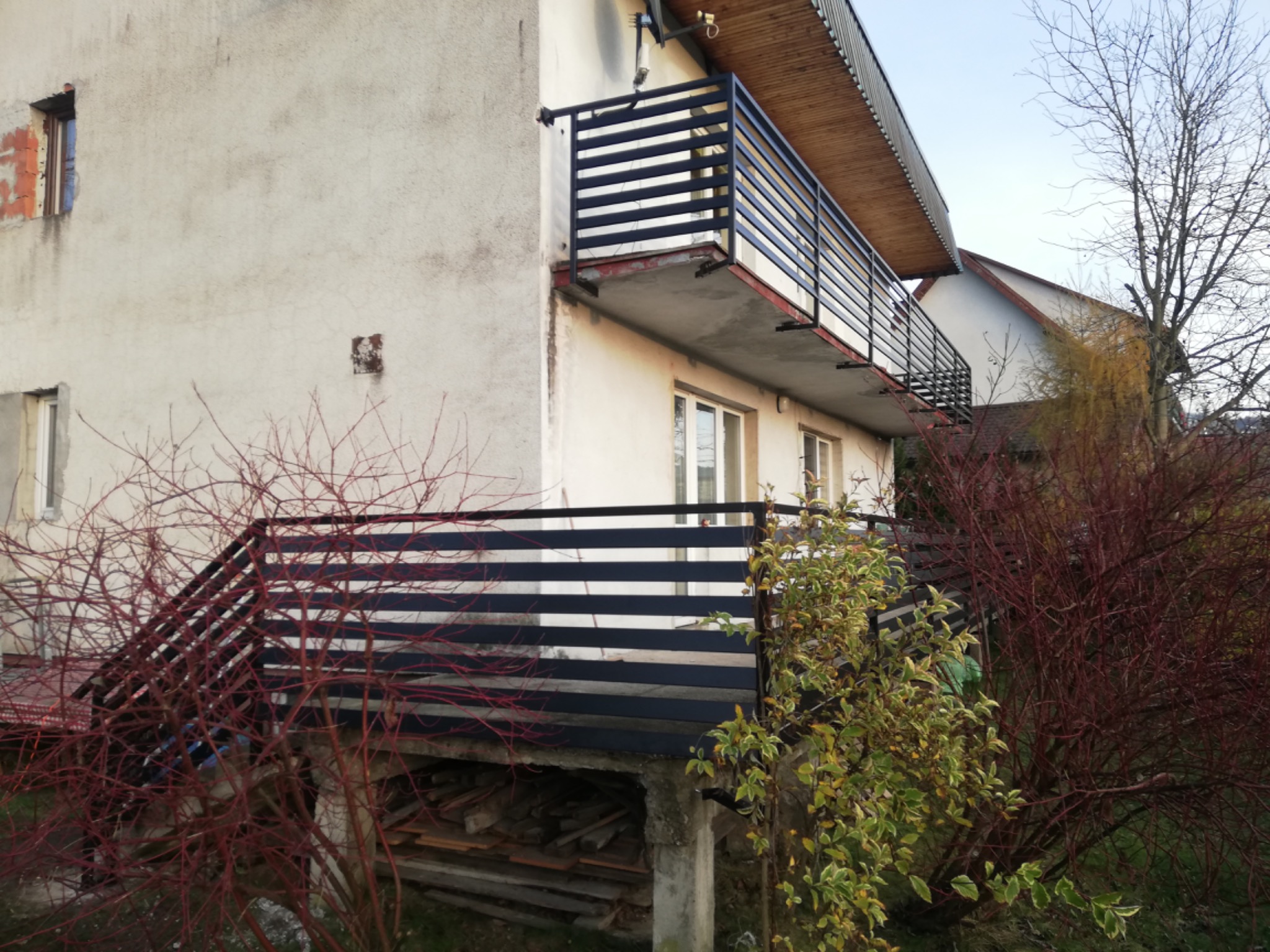 Dwupiętrowy dom z balkonami zabezpieczonymi poziomymi, czarnymi balustradami, widok na elewację z czerwonymi krzewami na pierwszym planie.