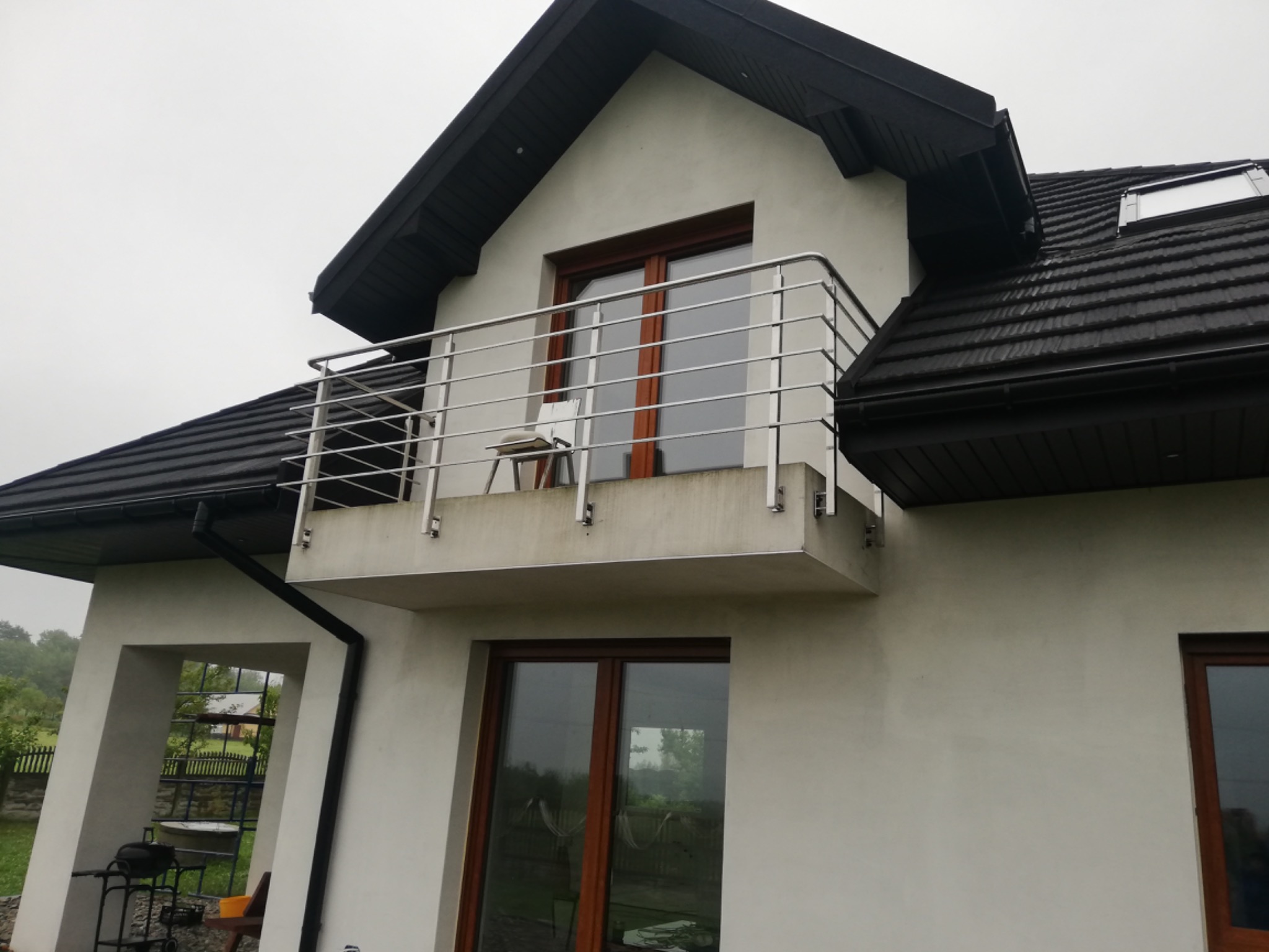 Dom z balkonem zabezpieczonym nowoczesną, poziomą balustradą nierdzewną z widocznym krzesłem na balkonie i oknem dachowym.