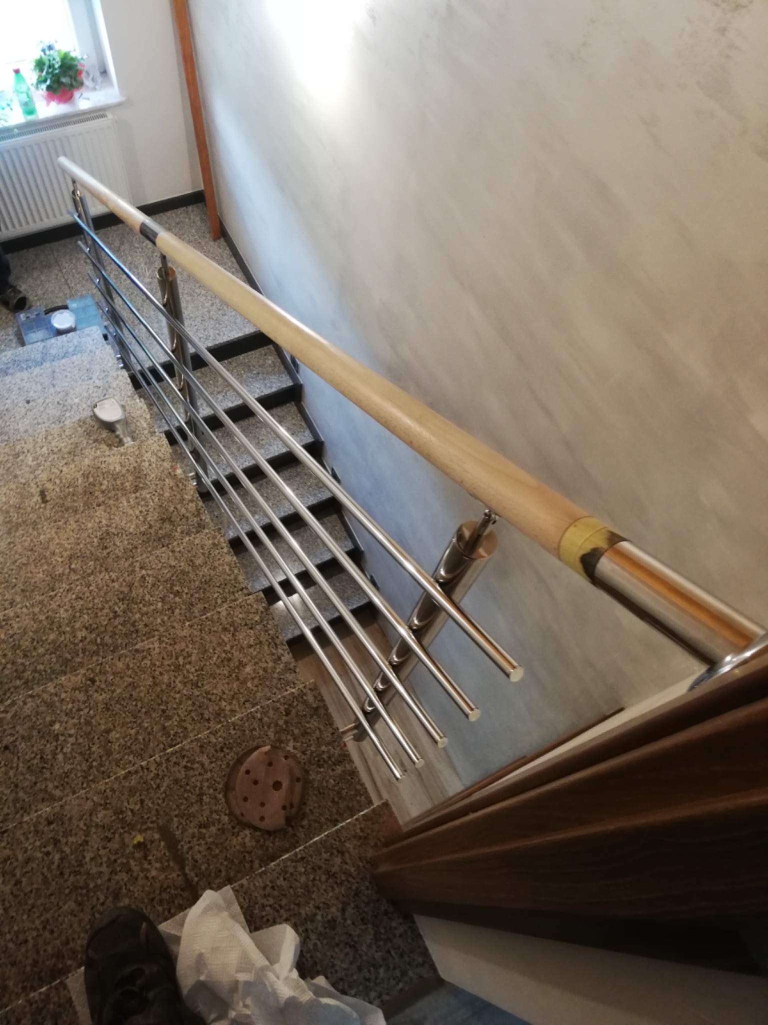 Wewnętrzna balustrada schodowa ze stali nierdzewnej z drewnianą poręczą, widok z góry na schody z terazzo i fragment podłogi z widocznymi śladami prac montażowych.