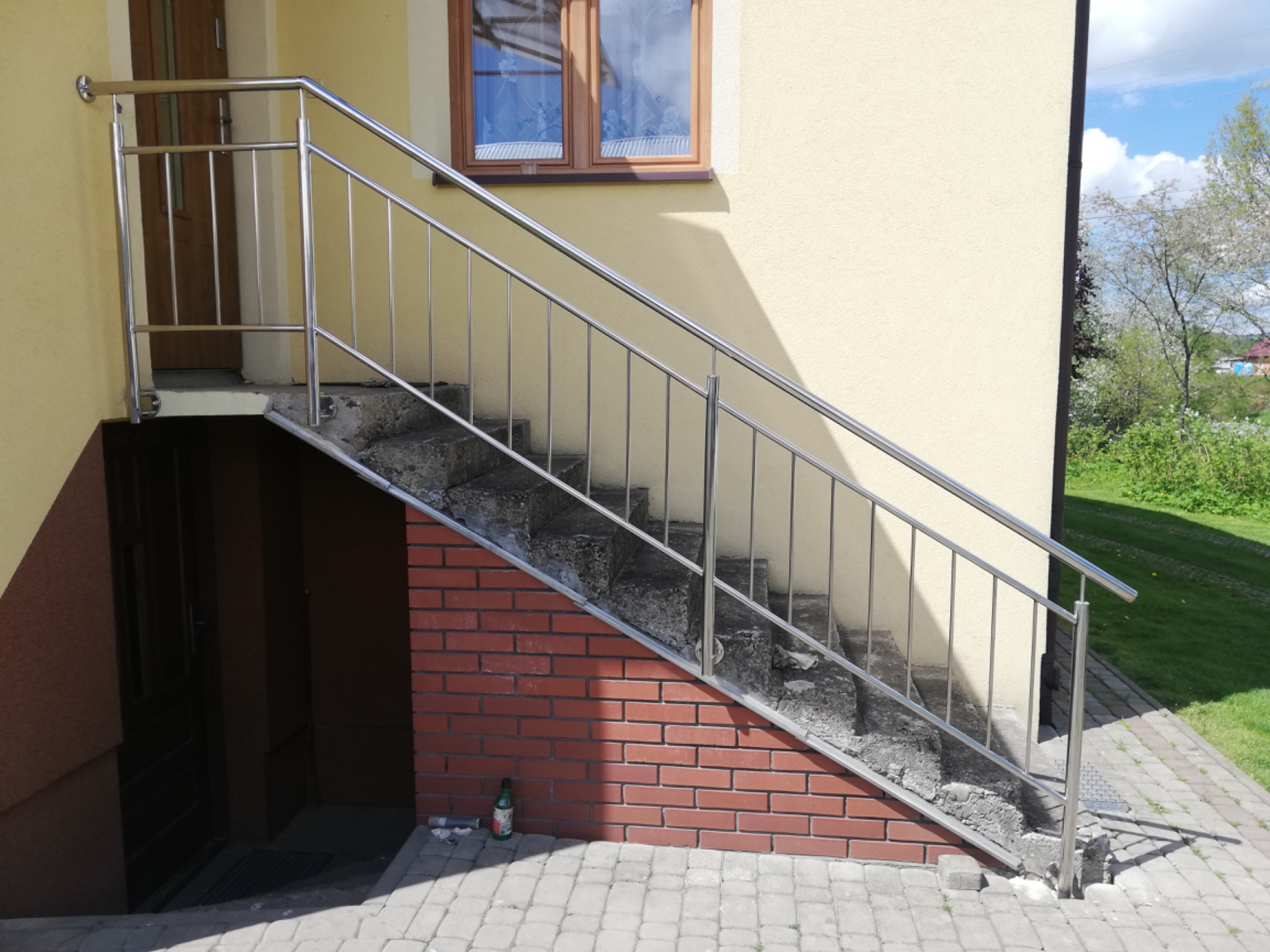 Zewnętrzne schody betonowe z prostą, metalową barierką przy wejściu do piwnicy, z widoczną butelką po napoju u podstawy ściany z cegły klinkierowej.