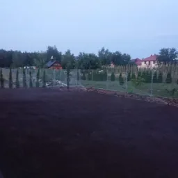 Przygotowany teren pod trawnik, świeżo wyrównana, ciemna gleba, obrzeża z kory, rząd tuj wzdłuż ogrodzenia z siatki, zabudowa jednorodzinna w tle, pochmurne niebo.