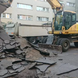 Ż&oacute;łta koparka PW 200 z pełną łyżką gruzu asfaltowego nad wywrotką na tle bloku mieszkalnego.