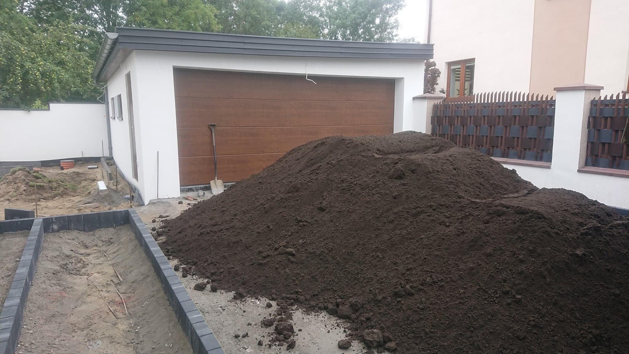 Kupa ciemnej ziemi przed garażem z brązową bramą, widoczna łopata, obrzeża chodnika w trakcie montażu, w tle ogrodzenie.